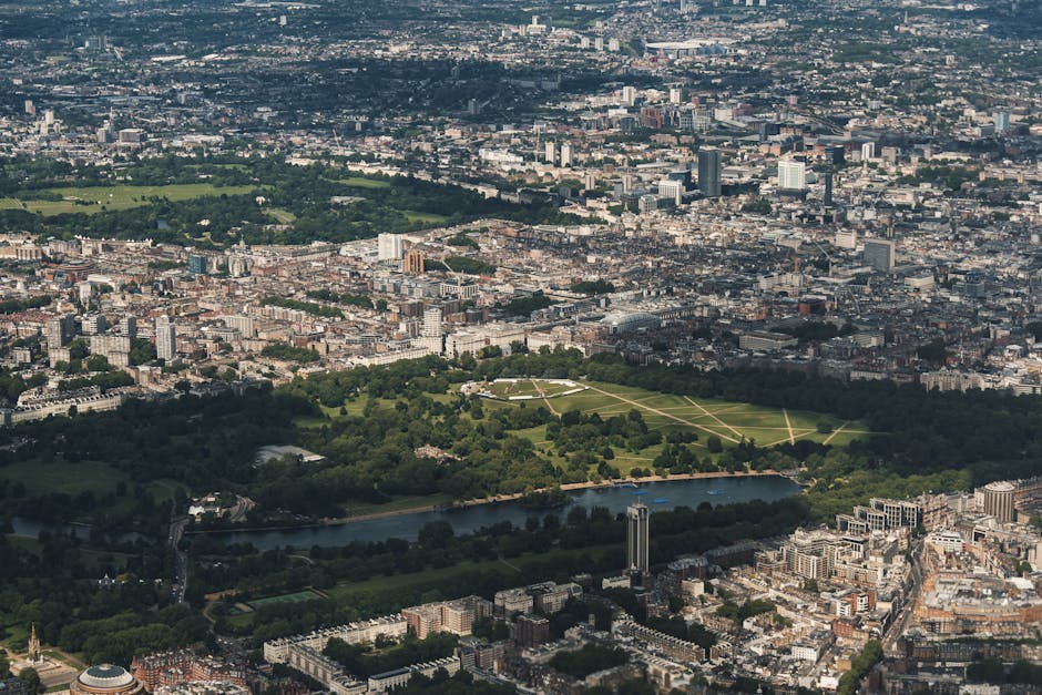 An aerial view of central London showing a large green park with pathways and a river running through it. Surrounding the park are densely packed urban areas with high-rise office buildings, residenti